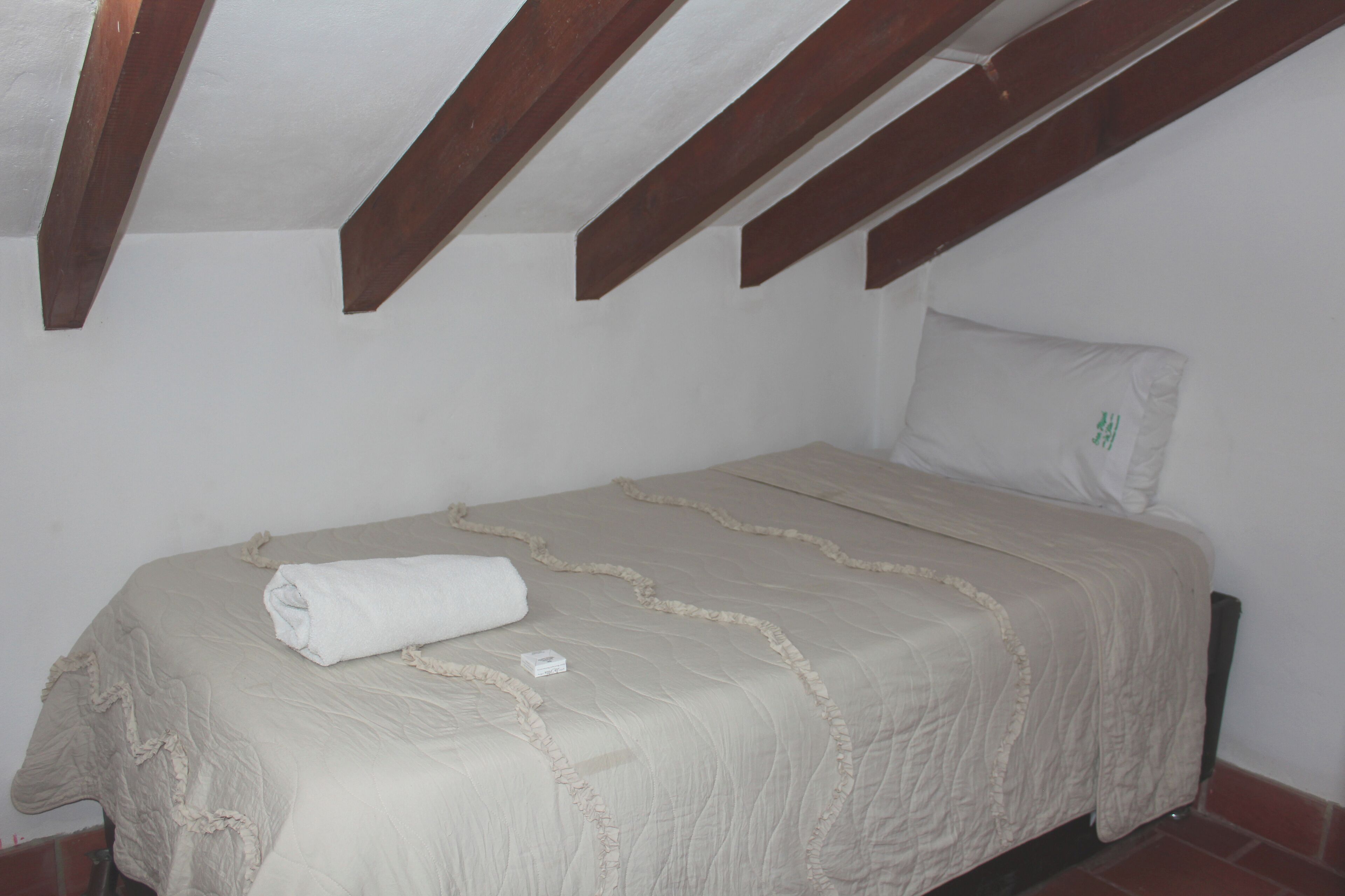 Un lit simple avec une couette beige et une serviette blanche roulée, des poutres en bois au plafond et un oreiller blanc orné d’un logo vert.