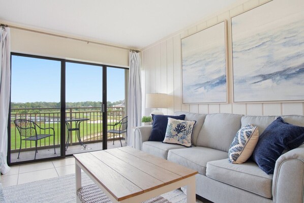 A living room with a sofa, coffee table, and a view of a golf course through a sliding glass door.