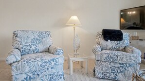 A living room with two patterned armchairs, a small white side table, a floor lamp, and a television on the wall.