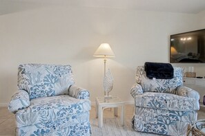 A living room with two patterned armchairs, a small white side table, a floor lamp, and a television on the wall.