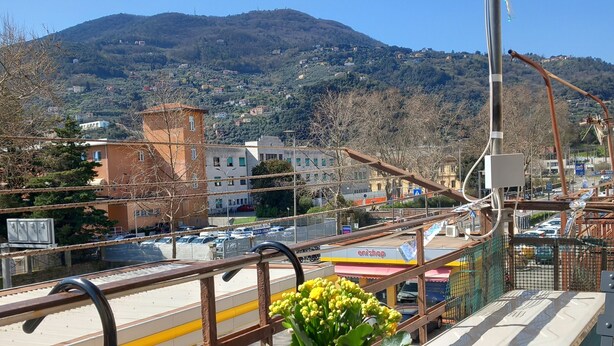 A balcony with a view of a hilly town, a yellow flower pot, and a gas station in the background.