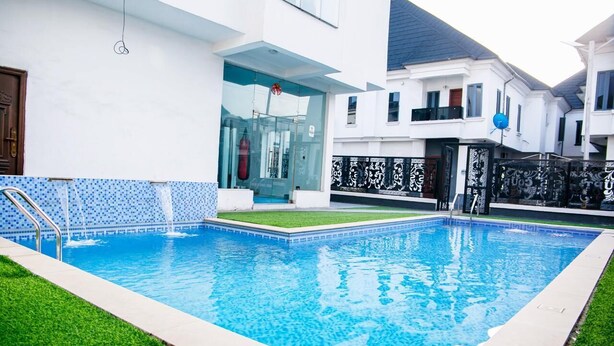 Una piscina con bordo piastrellato, un'area verdeggiante e una casa moderna con un balcone.
