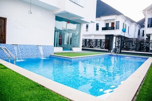 Een zwembad met betegelde rand, een gazon en een modern huis met balkon.