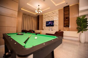 Une table de billard dans une pièce moderne, équipée d’un téléviseur, de sièges confortables et d’une grande plante.