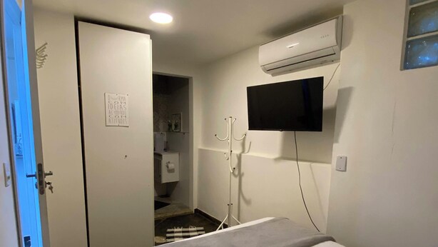 A hotel room with a bed, a TV mounted on the wall, an air conditioning unit, and a door leading to a bathroom. - Cozy Loft on Rua da Amizade - Itaipu (Itaipu Niterói)