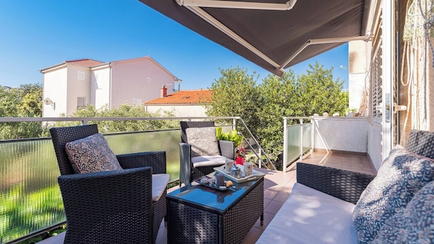 A balcony with wicker furniture, a table with a tray, and a view of a building and trees. - Apartment in Brodarica Near Beach & Krka Park (Sibenik)