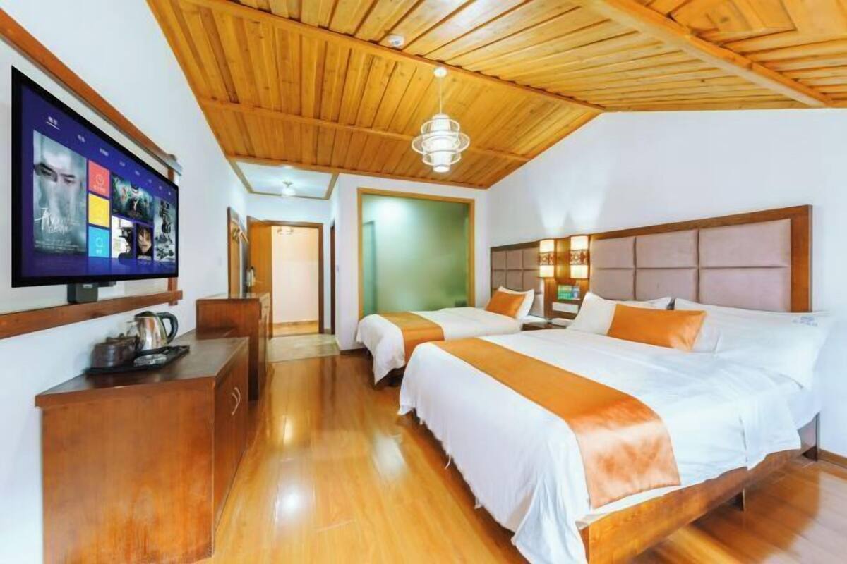 A hotel room with wooden flooring, a large bed with orange bedding, a smaller bed, a TV mounted on the wall, and a wooden ceiling.