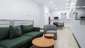 Una sala moderna con un sofá verde, un sillón gris y una mesa de centro de madera. Se observa una cocina al fondo con armarios blancos y un microondas.