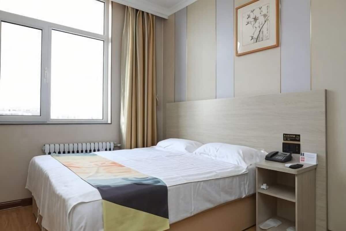 A hotel room with a bed, a bedside table with a phone and a lamp, a window with curtains, and a radiator.