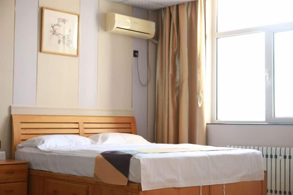 A bedroom with a wooden bed, a bedside table, an air conditioning unit, a radiator, and a window with curtains.