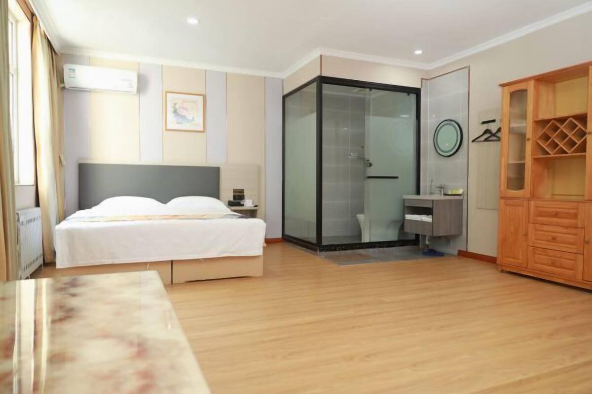 A hotel room with a bed, a glass shower enclosure, a wooden wardrobe, and a small table.