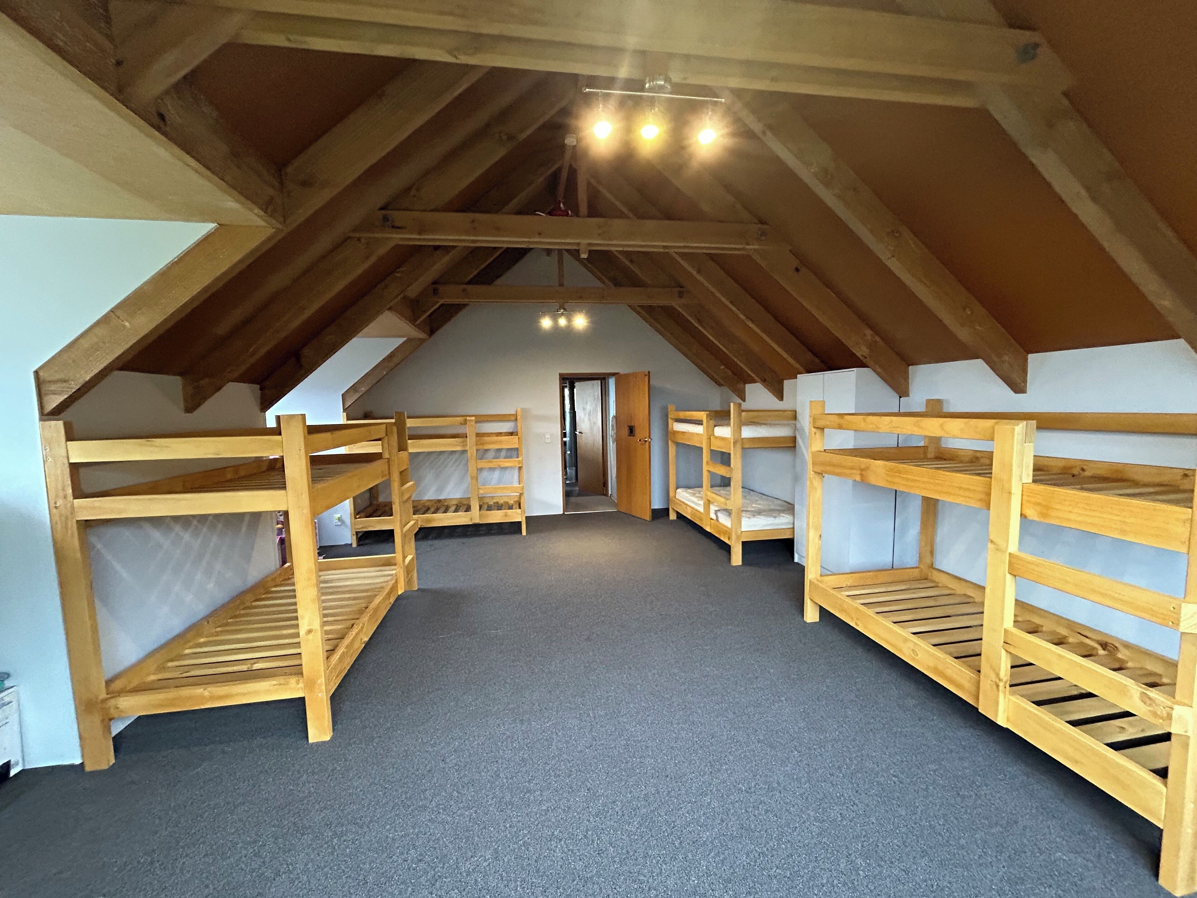 Une chambre de dortoir avec quatre lits superposés, un sol recouvert de moquette et un plafond en pente avec des poutres en bois.