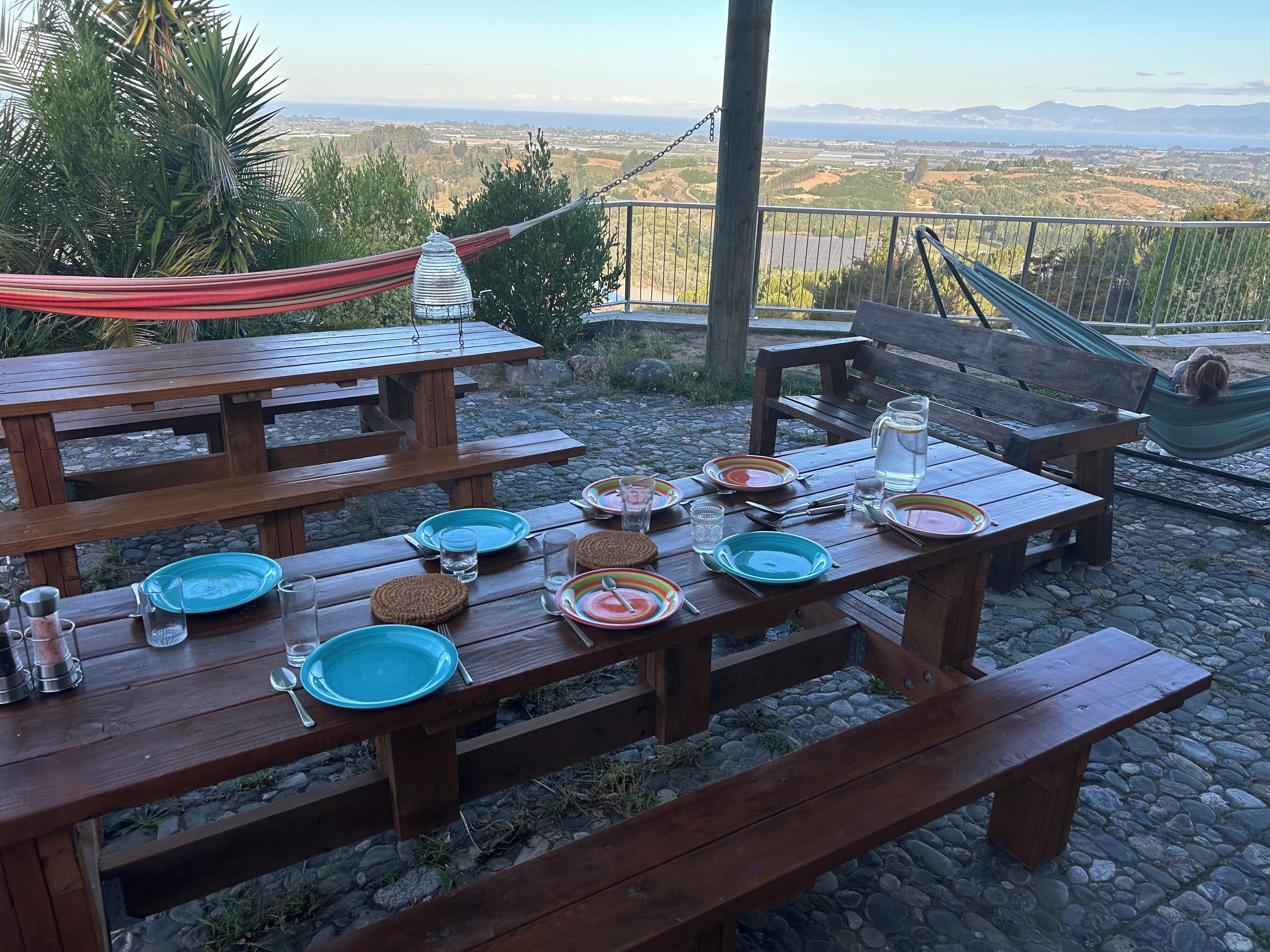 Un montaje para picnic con mesas y bancos de madera, rodeado de una vista panorámica de montañas y el mar.