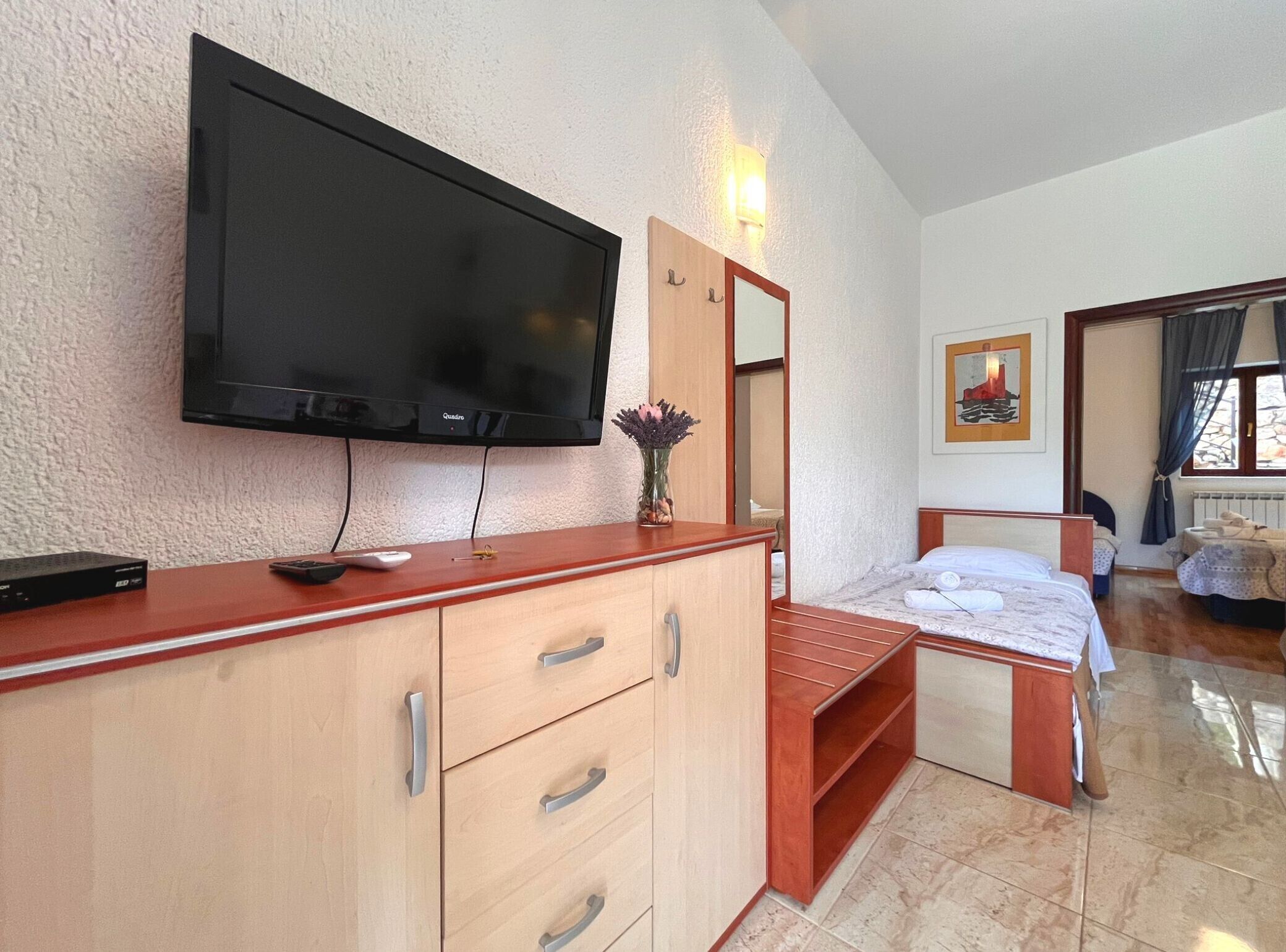 Ein Hotelzimmer mit Flachbildfernseher an der Wand, einer Holzkommode mit Schubladen, einem Bett mit Holz Kopfteil, einem Nachttisch und einem Fenster mit Vorhängen.