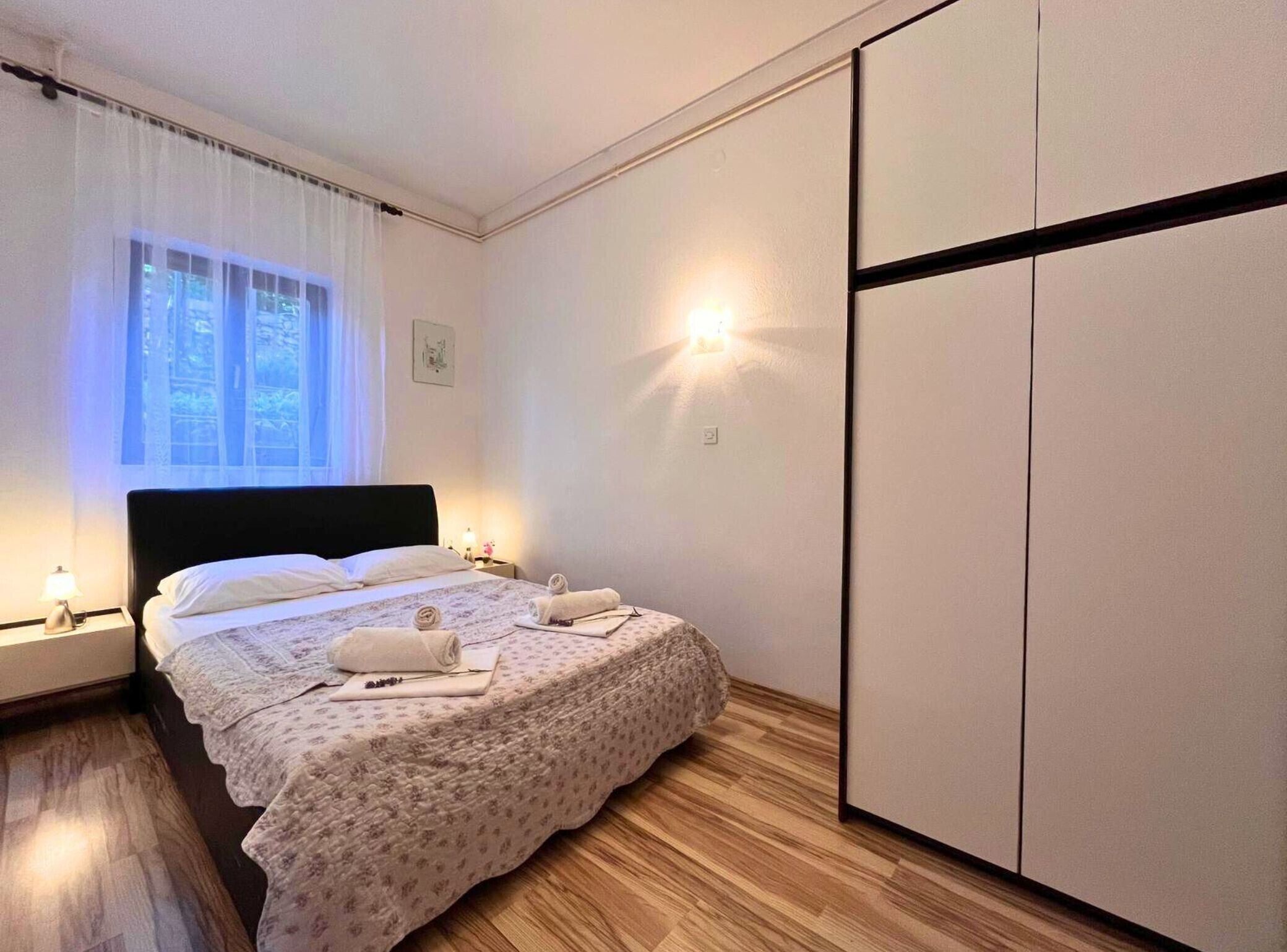 Un dormitorio con cama, lámparas de noche, armario y ventana con cortinas.