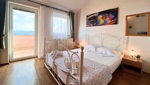 A bedroom with a bed, a nightstand, a painting, a window with curtains, and a view of the sea.