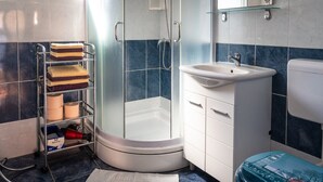 A modern bathroom with a glass-enclosed shower, a white sink, and a toilet. There is a towel rack with folded towels and a shelf with toilet paper and a red hairdryer.