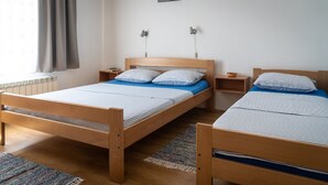 A room with two wooden beds, each with a blue mattress and striped bedding. There are bedside tables with lamps, a window with curtains, and a radiator.