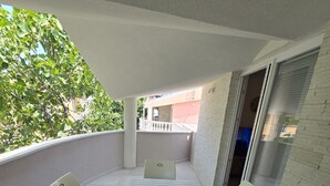 Un balcone con tavolo e sedie bianche, un oggetto blu sul tavolo e vista su un edificio e alberi.