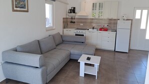 A compact living space with a grey sectional sofa, a small white table, a kitchenette with white cabinets, and a single window. - Nane Garden house with heated pool (Mirca)