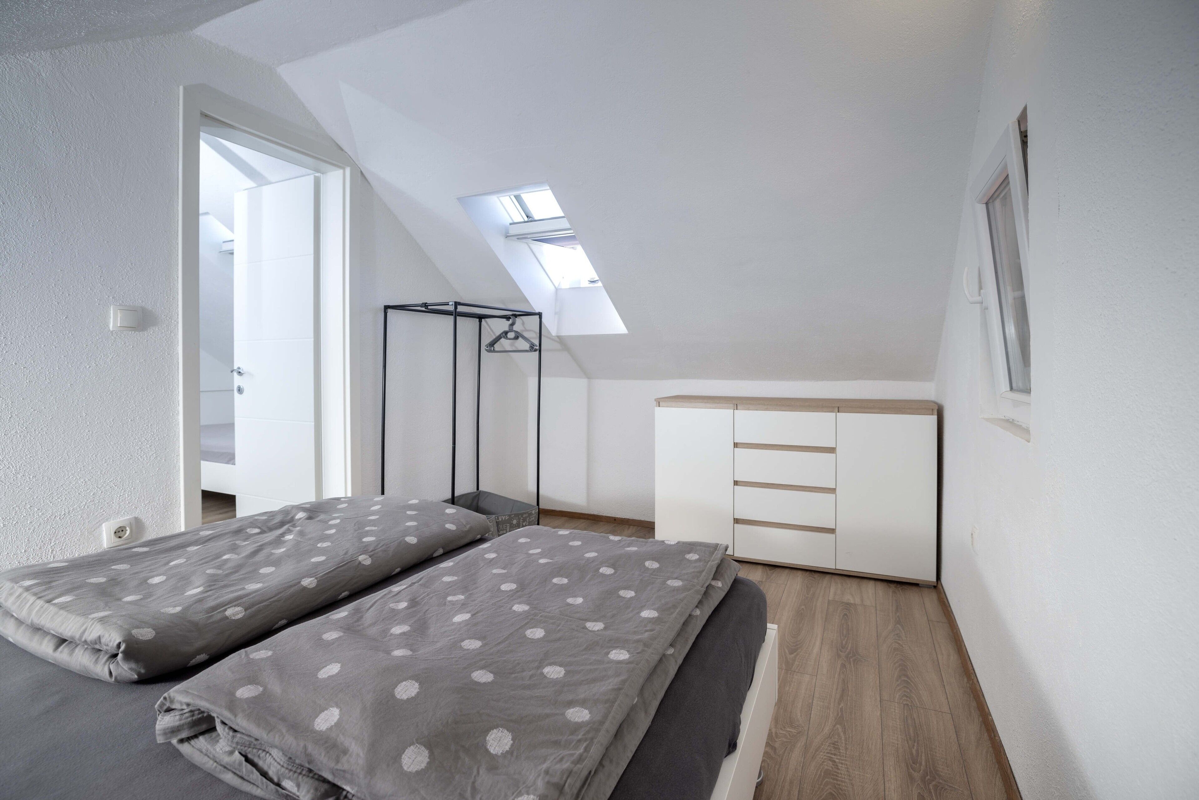 A bedroom with a sloped ceiling, a skylight, a bed with grey polka dot bedding, a white dresser, and a wooden floor.