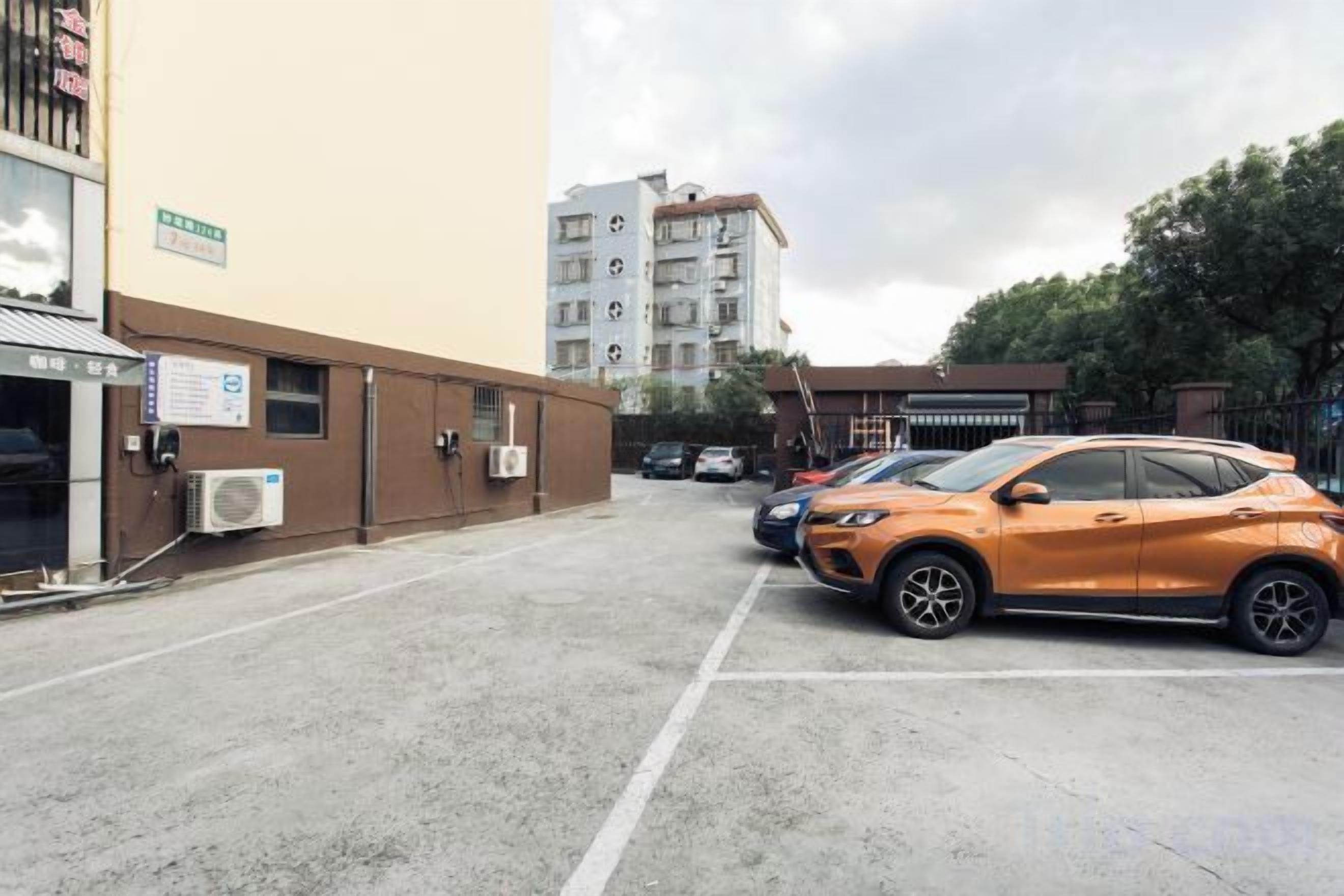 A parking lot with a building on the left and a multi-story structure in the background.