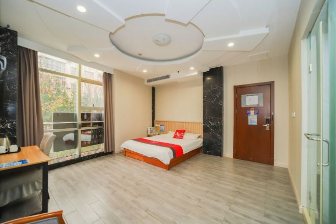 A hotel room with a bed, a desk, a window with curtains, a door, and a ceiling with recessed lighting.
