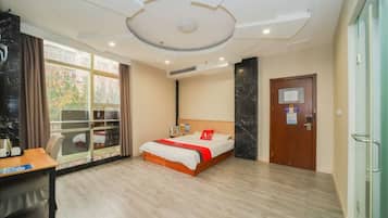 A hotel room with a bed, a desk, a window with curtains, a door, and a ceiling with recessed lighting.