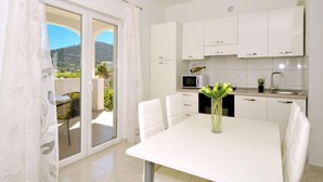 Une cuisine moderne avec des meubles blancs, une table à manger avec des chaises et une vue sur l’extérieur grâce à une porte vitrée.