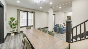 Una habitación amplia con una mesa de comedor de madera, sillas tapizadas en verde, una planta en maceta y una escalera que conduce a otra zona.