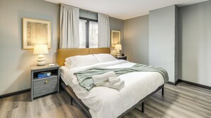 Una cama bien hecha con sábanas blancas, un plaid verde y una toalla doblada sobre un piso de madera. Se observa un mesita de noche con una lámpara y un libro.