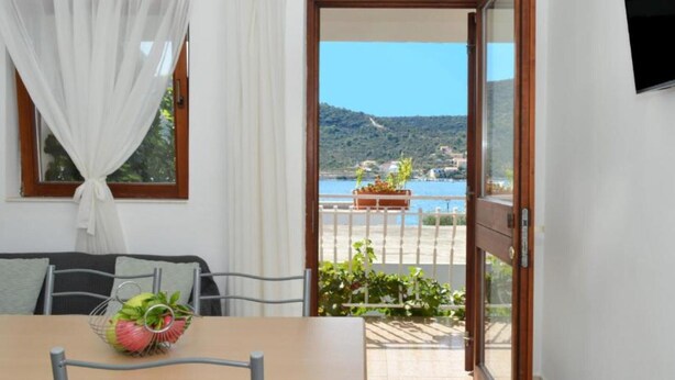 A room with a table, chairs, a window with curtains, and a view of a balcony with a railing and potted plants.
