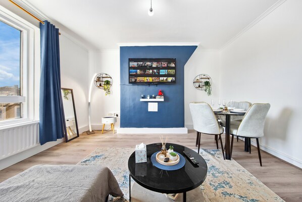 Un salon moderne avec un mur d’accent bleu, un téléviseur à écran plat, un coin repas comprenant une table et des chaises, et une grande fenêtre avec des rideaux.
