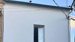 En tvåvåningsbyggnad med en vit fasad, en trädörr och två fönster med fönsterluckor. På bottenvåningen står två krukväxter.