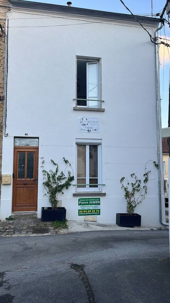 Um edifício de dois andares com fachada branca, porta de madeira e duas janelas com persianas. Há duas plantas em vasos no andar térreo.