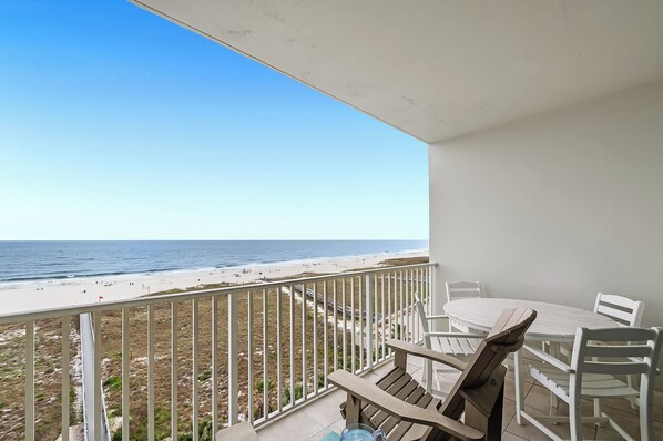 A balcony with a white table and chairs, overlooking a beach with a clear sky.