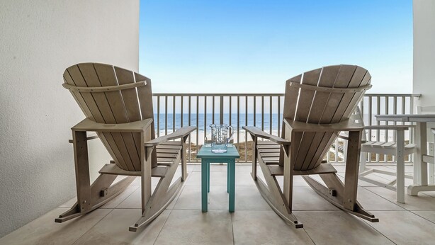 Un balcón con dos sillas Adirondack y una pequeña mesa, con vista a una playa.