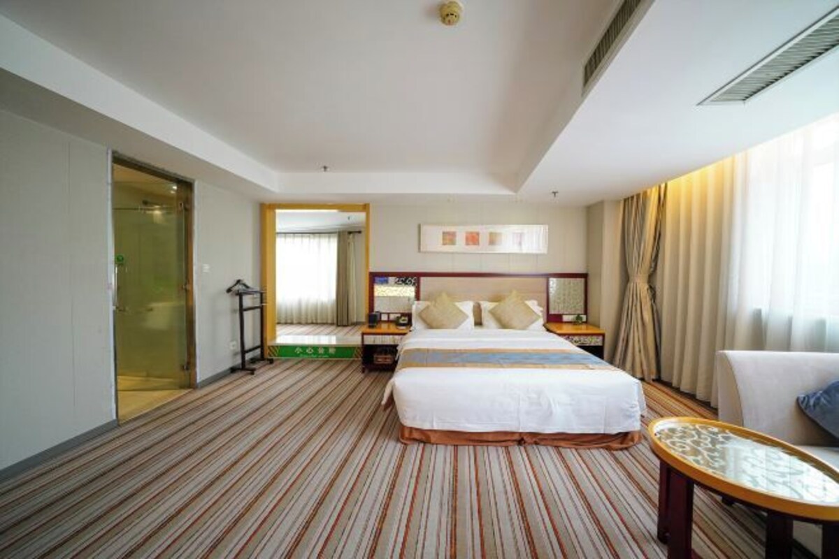 A hotel room with a large bed, a bedside table, a chair, and a view of the hallway.