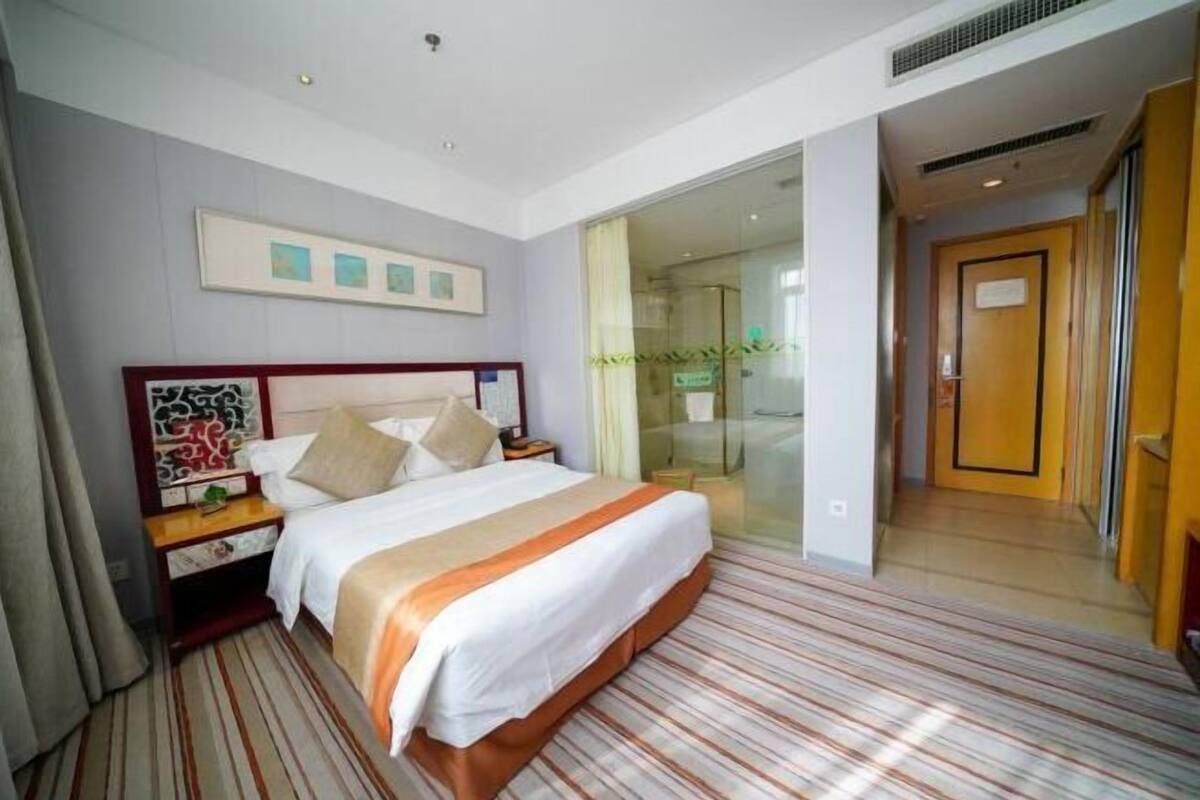 A hotel room with a bed, a nightstand, a glass door leading to a hallway, and a framed picture on the wall.