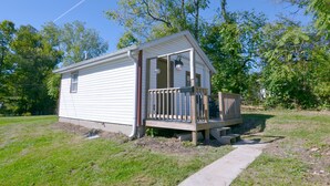 A small, single-story cabin with a front porch and a deck, surrounded by trees and a clear sky.
