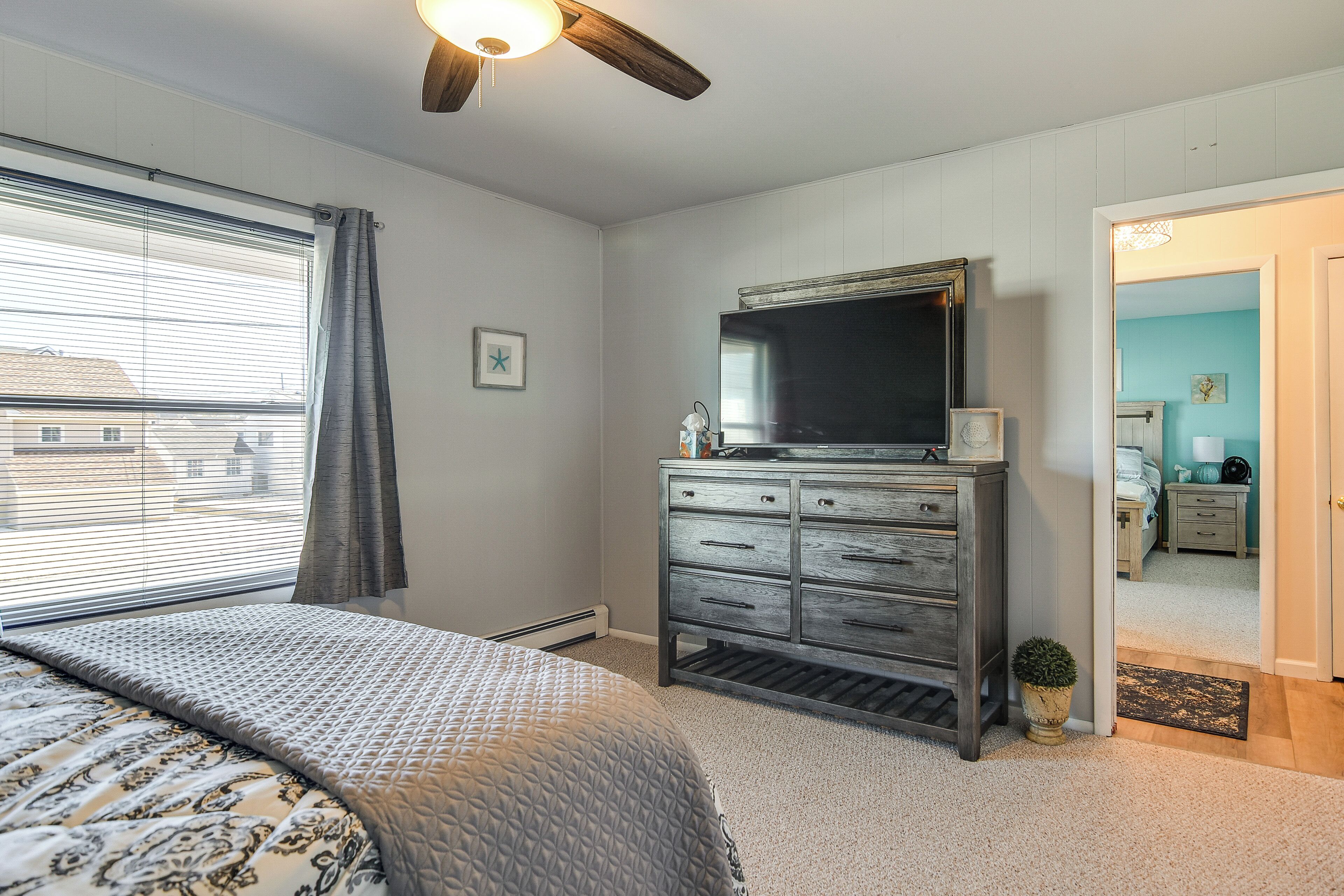 A bedroom with a bed, a dresser, a ceiling fan, a TV, and a window with blinds.
