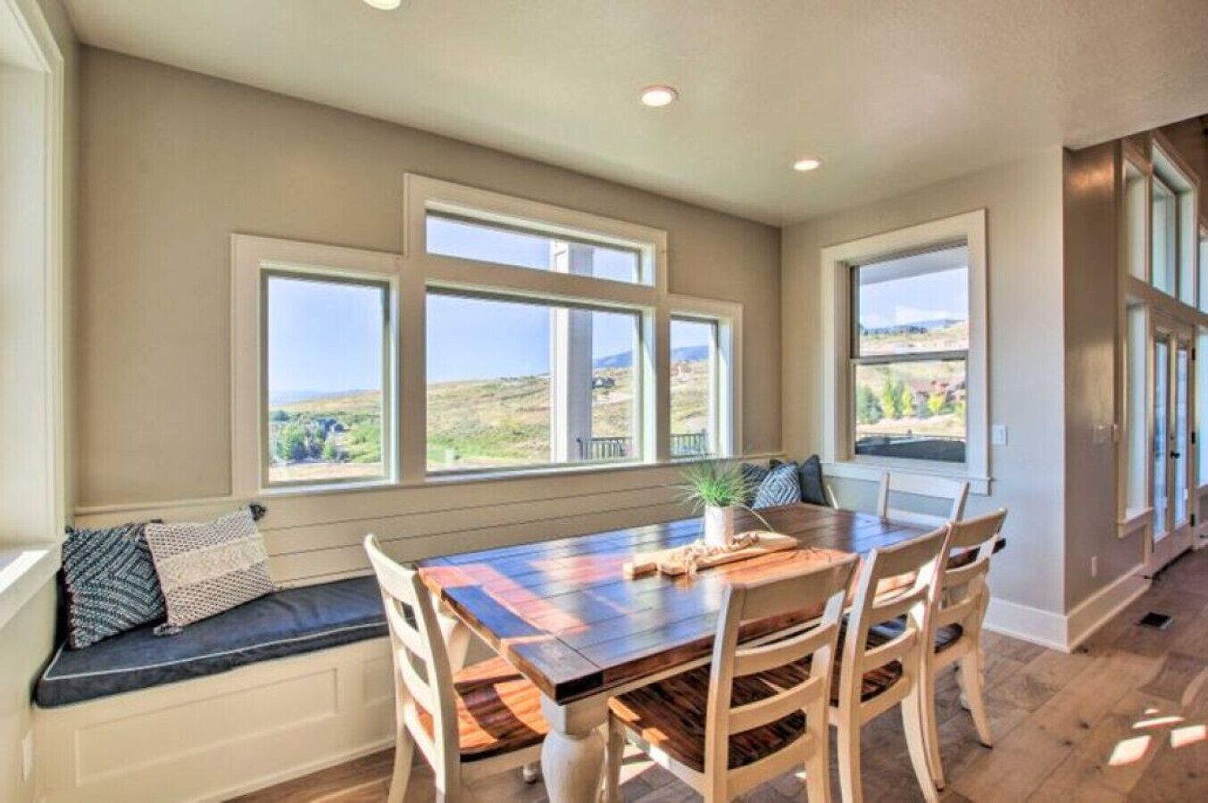 A dining area with a wooden table and chairs, large windows with views of the outdoors, and a built-in bench with cushions.