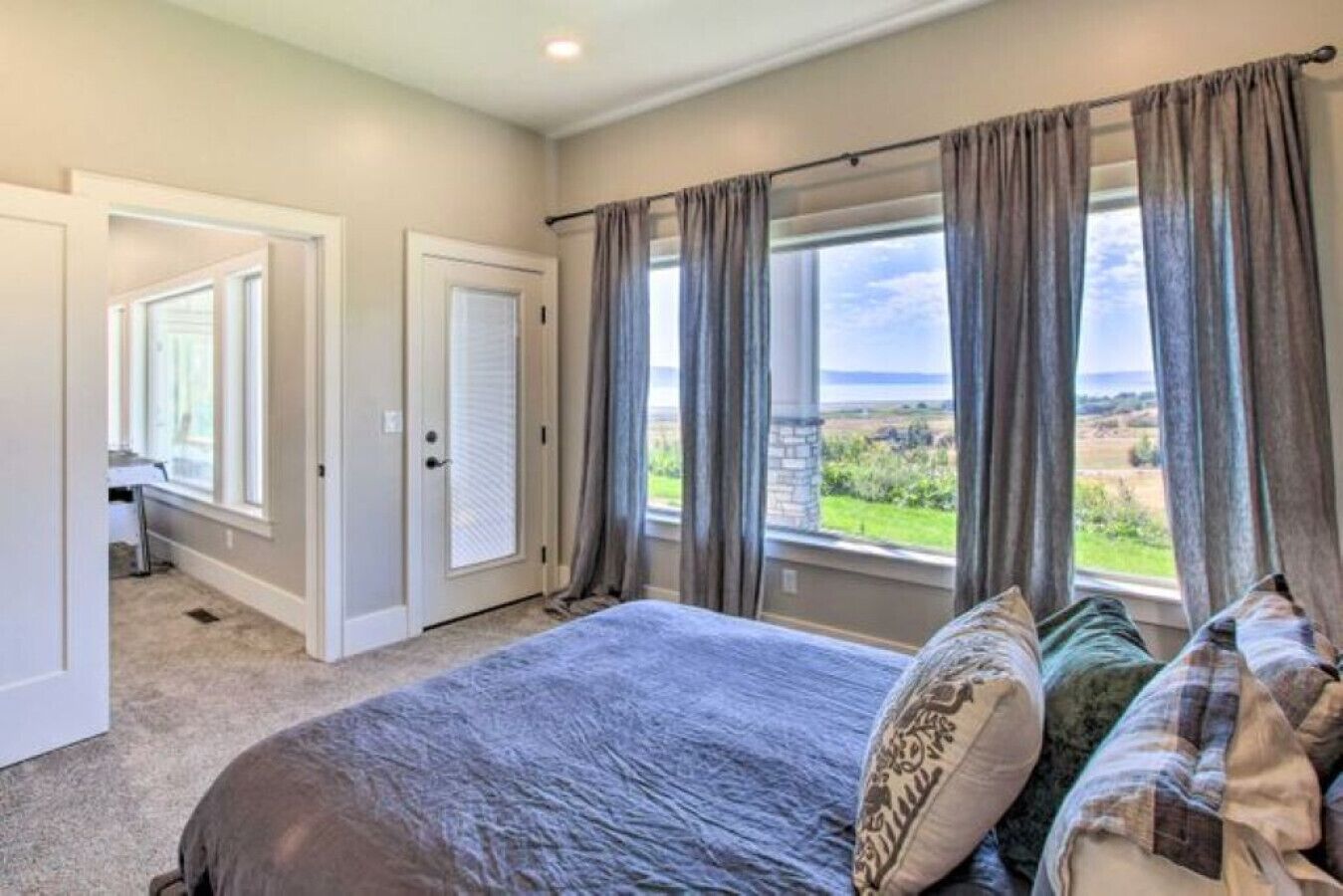 A bedroom with a large bed, two windows with curtains, a door, and a view of the outdoors.