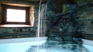 A rustic stone interior with a bathtub, a window, and a waterfall feature.