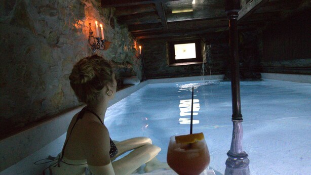 A person sitting by a pool with candles on the wall, a window, and a drink on the table.