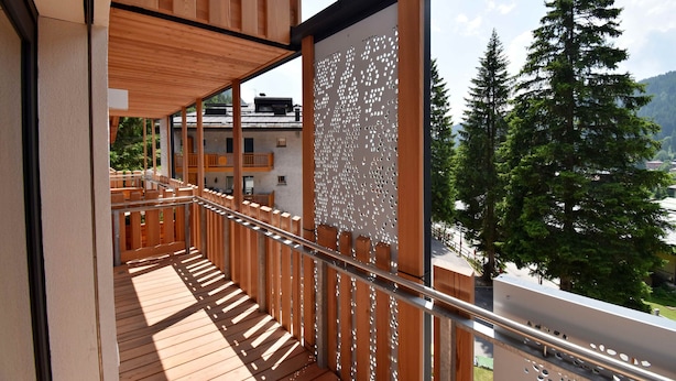 Ein Holzbalkon mit Metallgeländer, mit Blick auf eine gebirgige Landschaft mit Bäumen und Gebäuden.