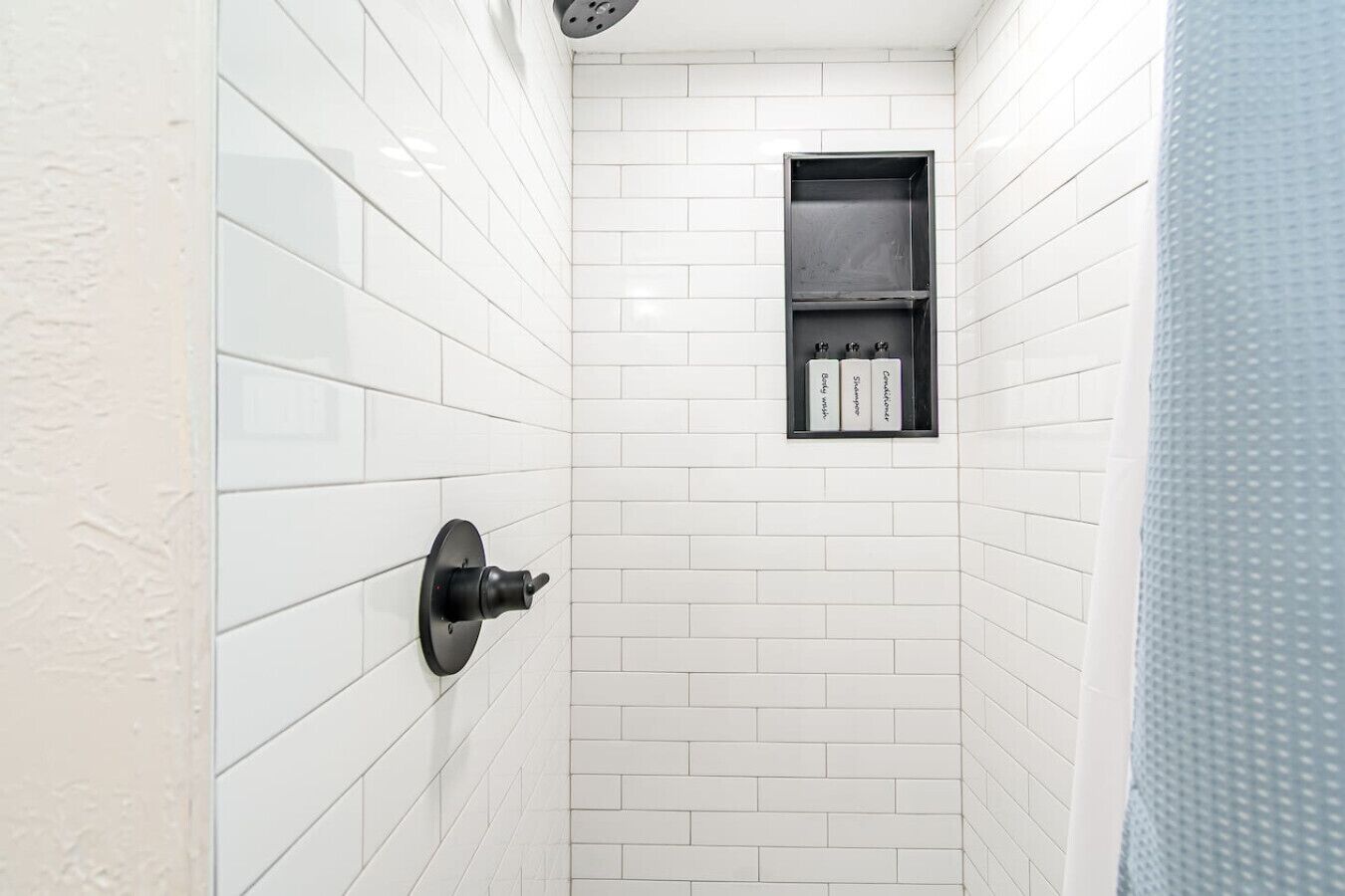 Um chuveiro pequeno revestido de azulejos brancos, com um dispensador de toalhas preto e uma prateleira acima.