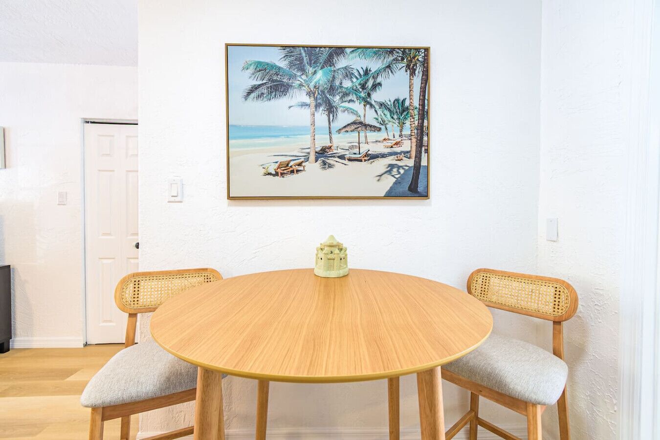 A round wooden table with two wicker chairs, a small decorative item on the table, and a beach scene painting on the wall.