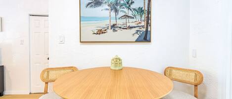 A round wooden table with two wicker chairs, a small decorative item on the table, and a beach scene painting on the wall.