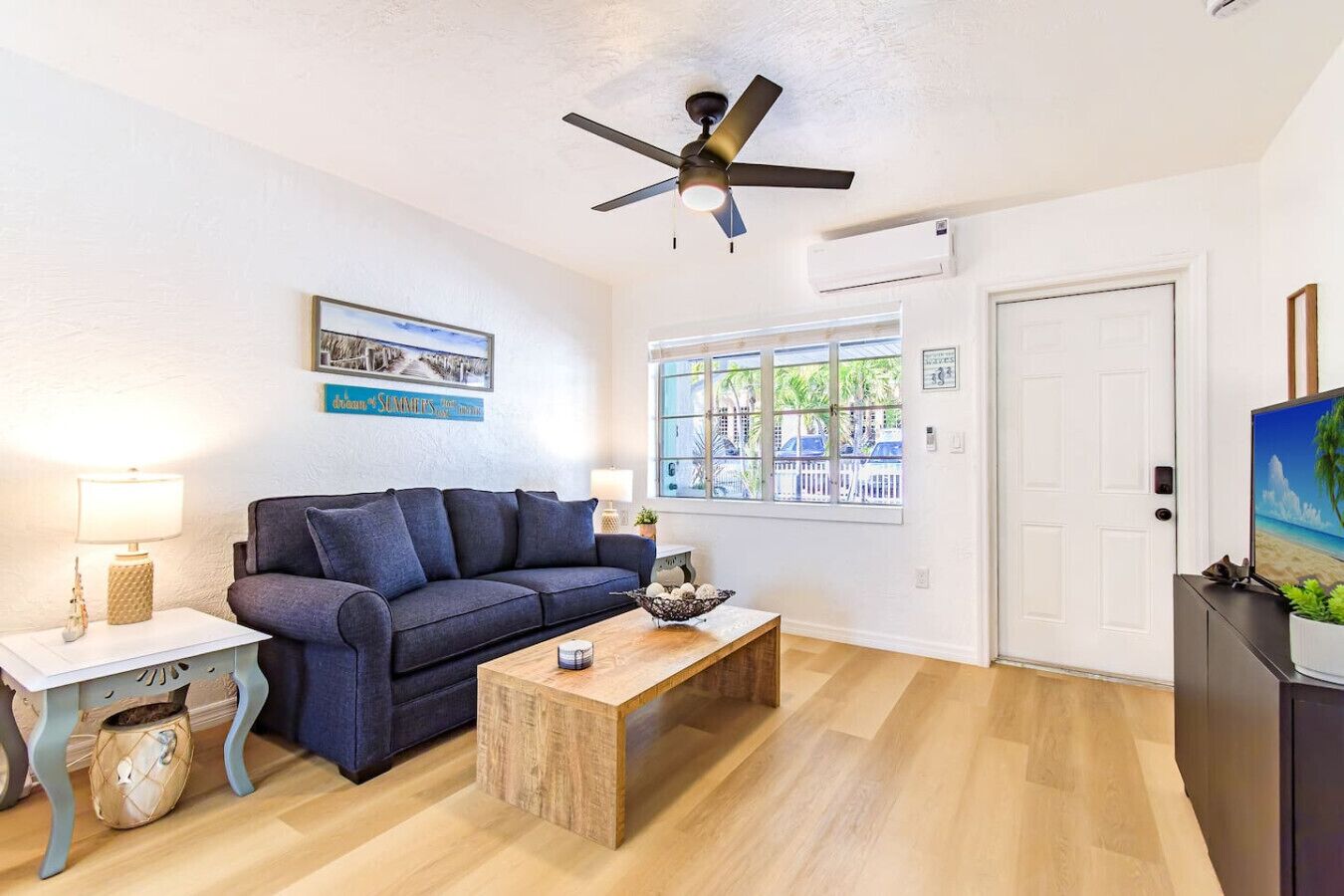 A living room with a dark blue sofa, a wooden coffee table, a ceiling fan, a window with a view of palm trees, and a door.